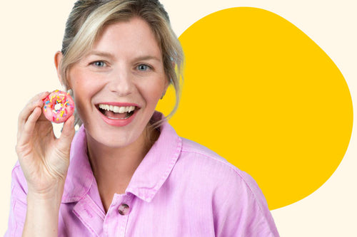 A blonde woman in a pink shirt smiling with a pink dipped donut in her hand